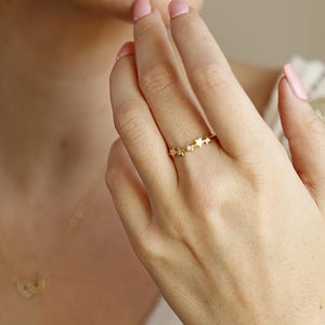 A person's hand wearing a gold ring with five diamond-studded stars, with a blurred gold necklace visible.