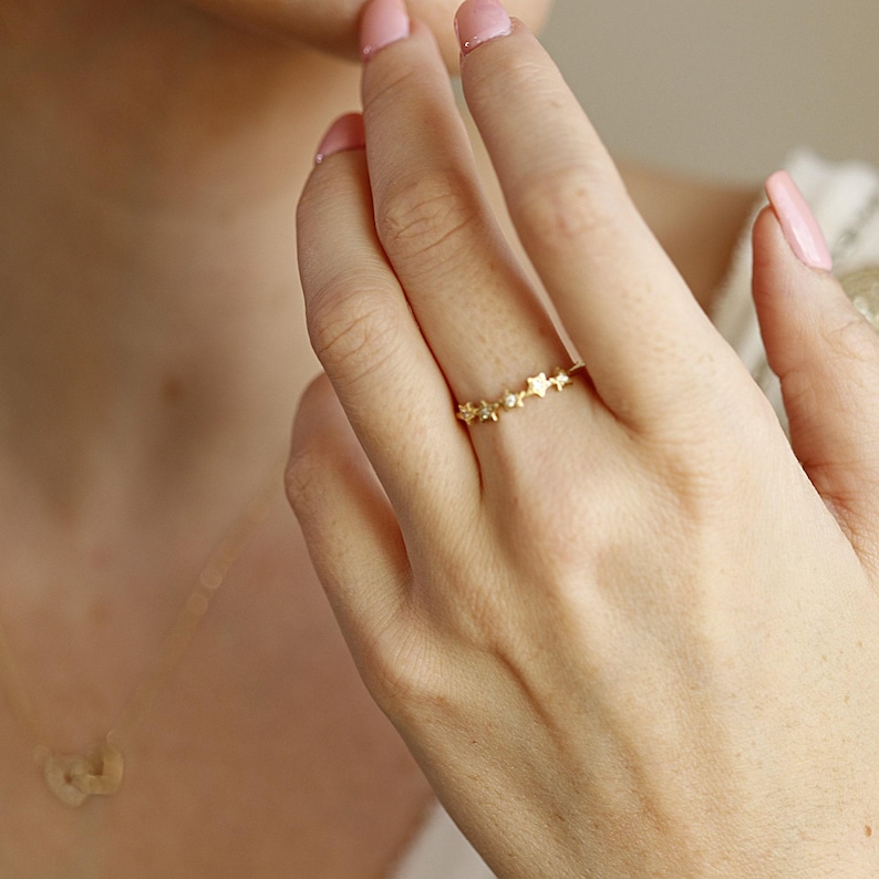A person's hand wearing a gold ring with five diamond-studded stars, with a blurred gold necklace visible.