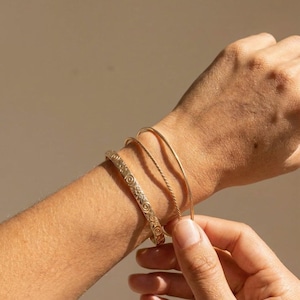 A person's arm wearing three gold bracelets: one engraved with a floral pattern, and two plain, thin bangles.