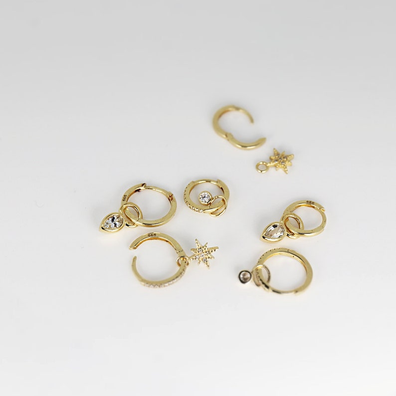 May include: A set of gold hoop earrings with various charms, including a star, a teardrop, and a round stone. The earrings are on a white background.