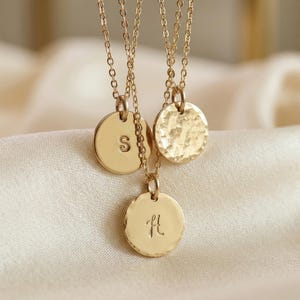 May include: Three gold-colored pendant necklaces. One pendant has the letter "S" engraved, another has a hammered texture, and the third has the letter "H" engraved. The necklaces hang from delicate gold chains.