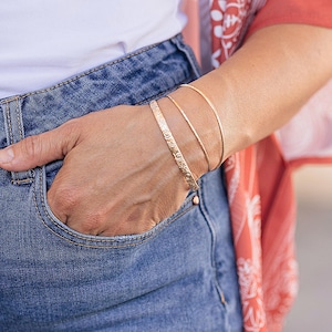 A person's arm wearing three rose gold bracelets: one engraved with a floral pattern, and two plain, thin bangles, with a blue jean pocket visible.