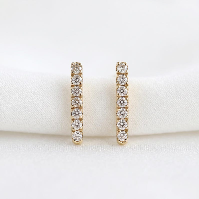 Two vertical gold diamond-pave bar earrings placed upright on a white fabric surface.