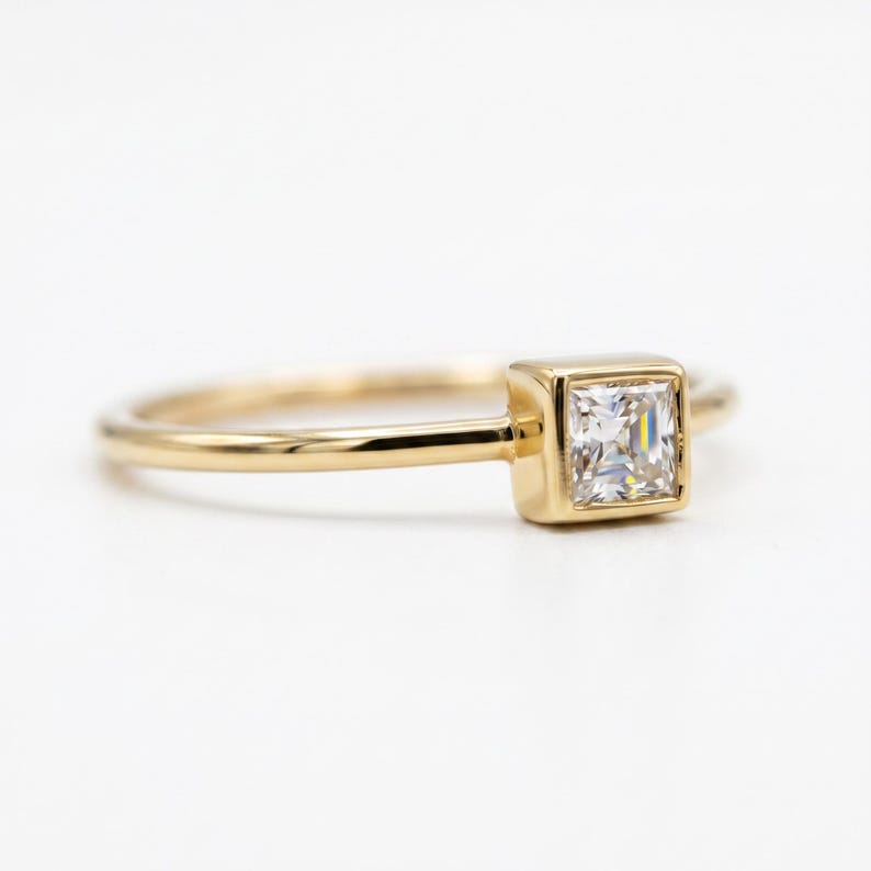A side profile view of a thin gold band with a bezel-set square princess cut diamond against a plain white background.