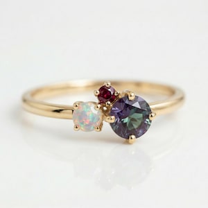May include: A gold ring featuring three gemstones: a large, round, deep teal stone, an opal, and a small, round, red stone. The ring has a delicate band and is set against a white background.