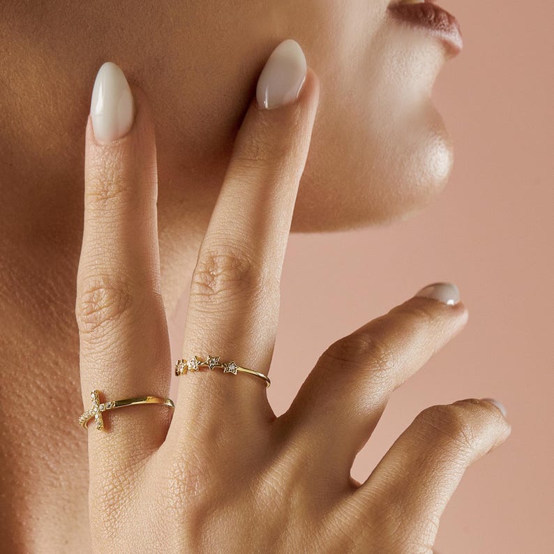 A person's hand with two gold rings: one with a diamond-studded cross and another with five diamond-studded stars.