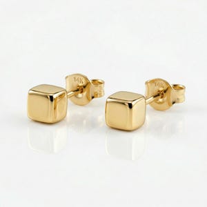 May include: A pair of gold cube stud earrings. Each earring features a small, polished gold cube with a post and butterfly backing. The earrings are set against a white background, highlighting their reflective surface and simple design.