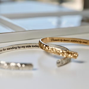 Two open cuff bracelets, one silver and one gold, with engraved text on the inside that reads: "Thank you for standing by my side today and always" and "Friends by heart, sisters by soul."