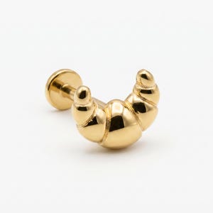 May include: A gold-colored earring in the shape of a croissant. The earring has a flat back and a curved, detailed front, with a shiny, reflective surface. The design is a miniature, stylized representation of the baked good.