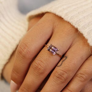 A hand wearing a yellow gold ring with an emerald-cut ametrine stone, positioned near a white knitted sleeve.