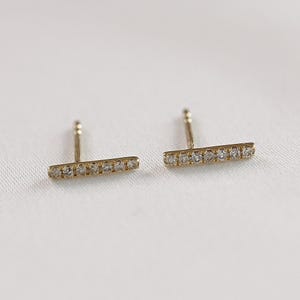 Two horizontal gold bar stud earrings with embedded pavé diamonds resting on a neutral fabric.