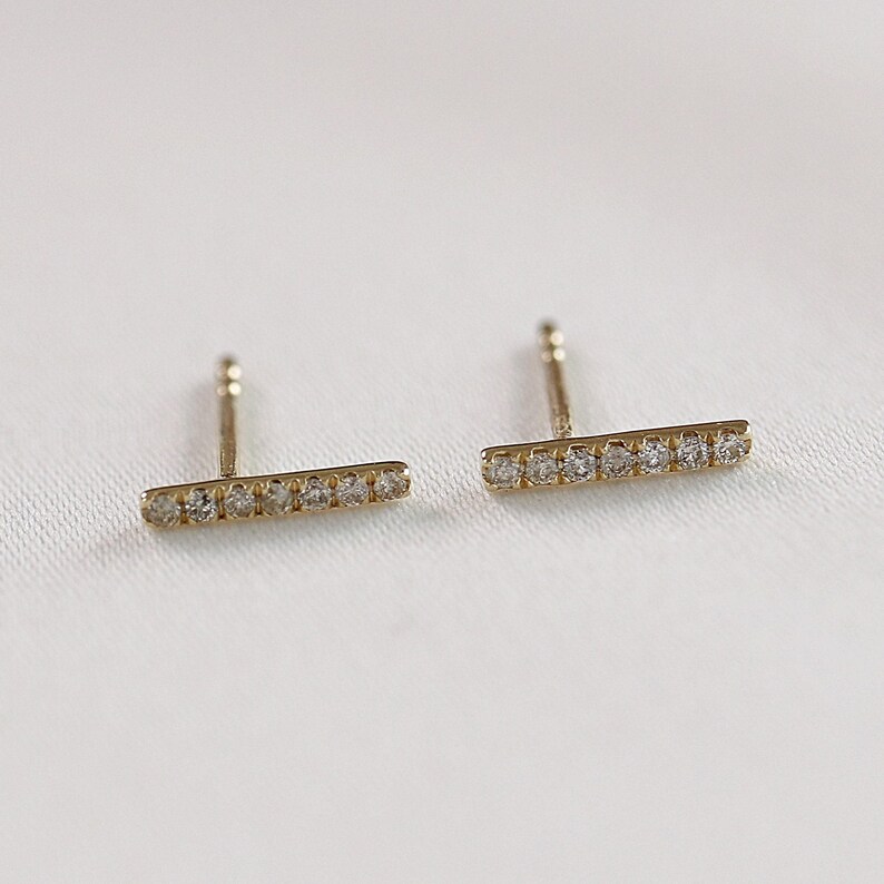 Two horizontal gold bar stud earrings with embedded pavé diamonds resting on a neutral fabric.