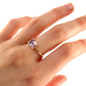 A hand wearing a thin yellow gold ring set with an emerald-cut purple gemstone against a white backdrop.