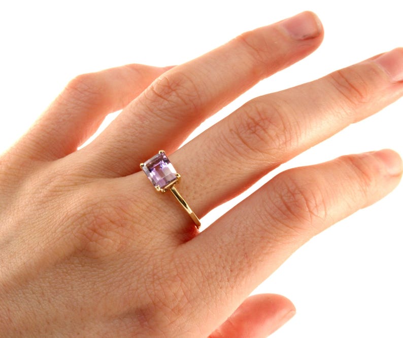 A hand wearing a thin yellow gold ring set with an emerald-cut purple gemstone against a white backdrop.
