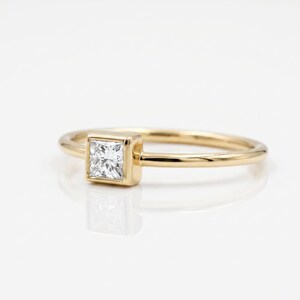 A thin gold ring featuring a bezel-set princess cut diamond viewed from an angled perspective against a white background.