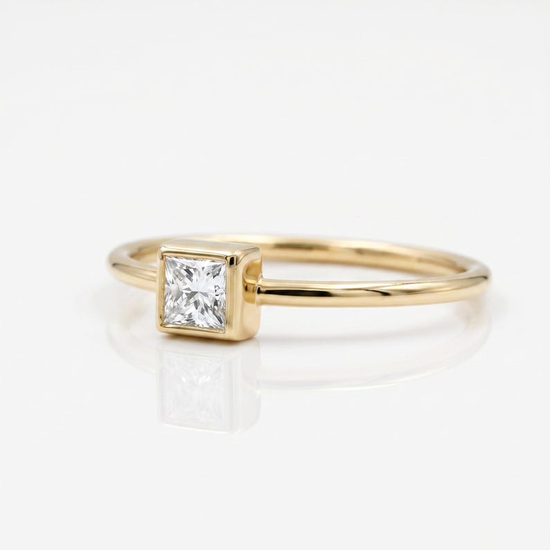 A thin gold ring featuring a bezel-set princess cut diamond viewed from an angled perspective against a white background.