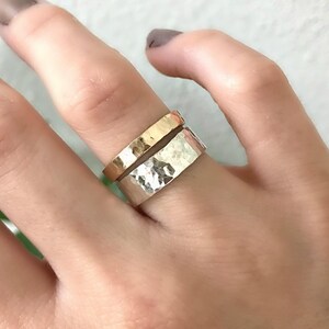 A wide, hammered silver band ring and a narrower, hammered gold band ring are stacked on a finger, with green leaves in the blurred background.