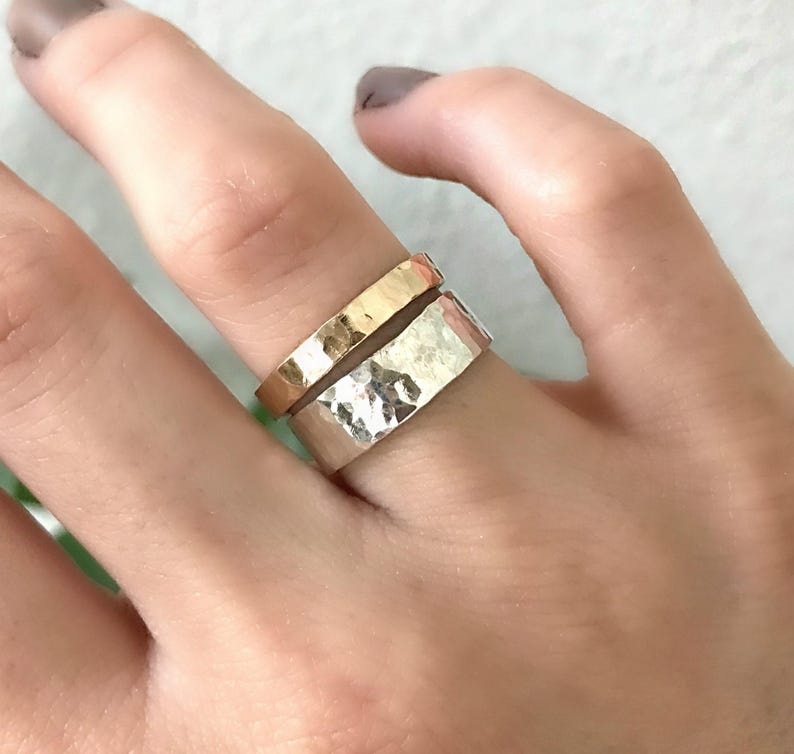 A wide, hammered silver band ring and a narrower, hammered gold band ring are stacked on a finger, with green leaves in the blurred background.