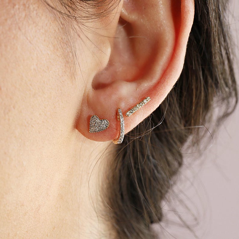 Side view of an ear wearing multiple gold and diamond earrings including a heart stud, a small hoop, and a bar stud.