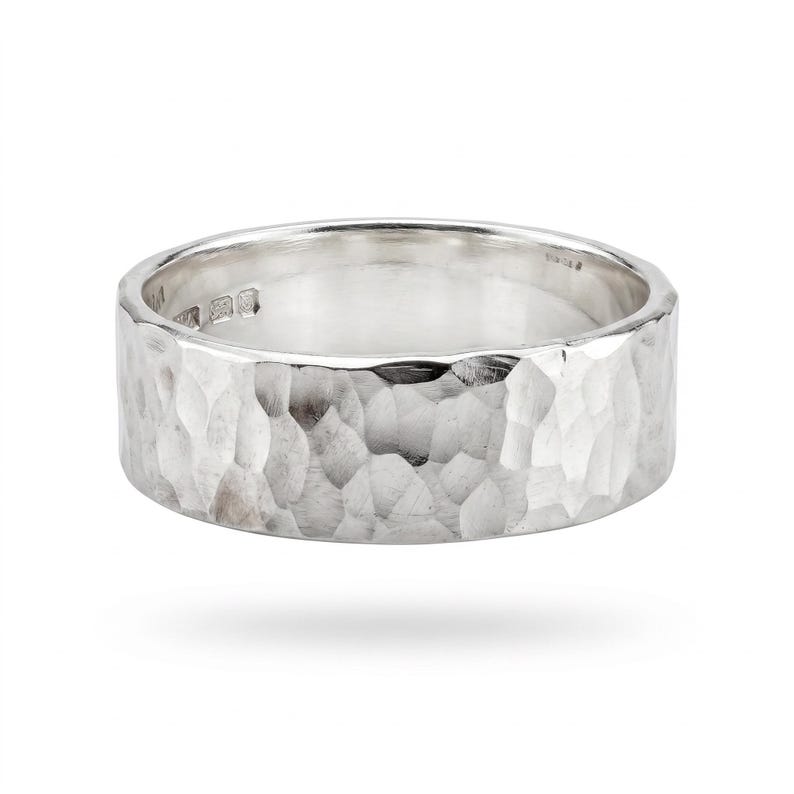 A single, wide, hammered silver band ring with a slightly irregular edge is shown on a white background, casting a shadow.