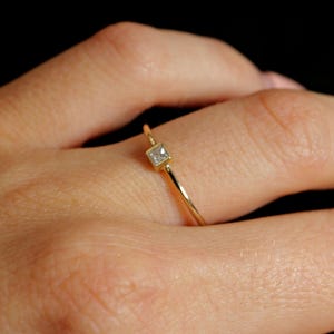 A close-up of a hand wearing a dainty gold ring with a small square-set diamond against a dark background.