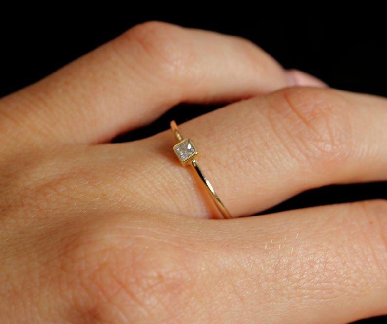 A close-up of a hand wearing a dainty gold ring with a small square-set diamond against a dark background.