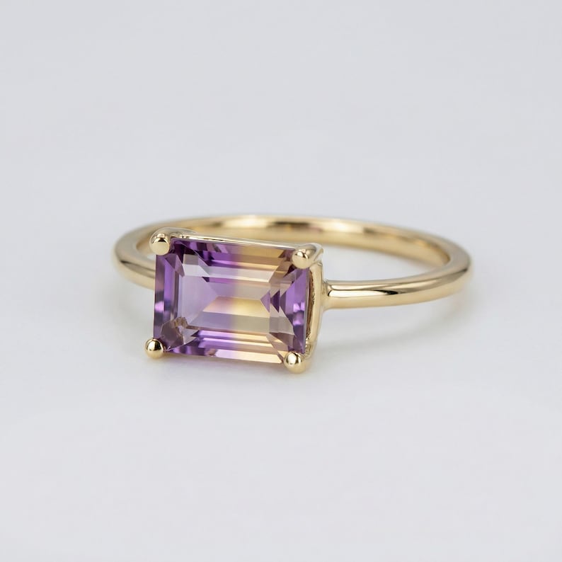 A close-up of a handcrafted ring with a large, rectangular emerald-cut ametrine stone. The gemstone shows a beautiful gradient of deep purple and honey yellow, set in a sleek, minimalist metal setting on a white background.