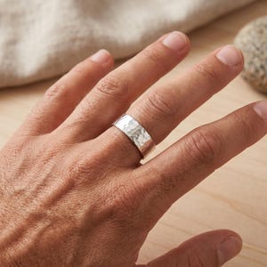 A wide, hammered silver band ring is worn on the ring finger of a person's hand, with a blurred background of a light cloth and a stone.