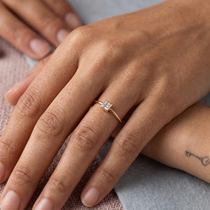 A hand wearing a thin gold ring with a single square stone on the ring finger.