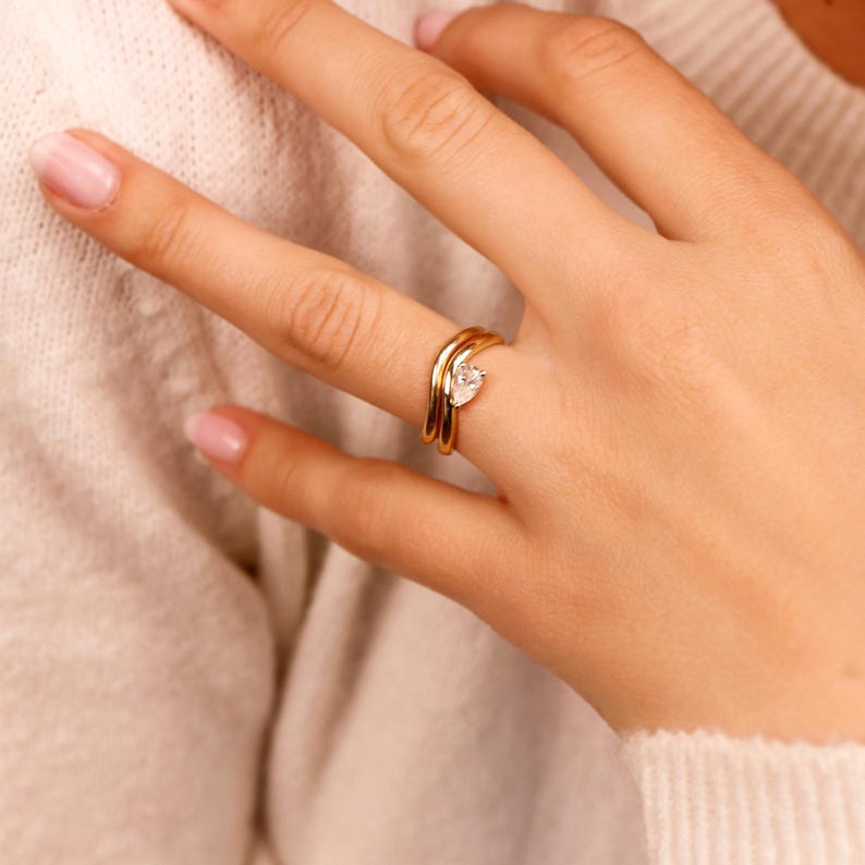 Woman wearing a pear-shaped moissanite ring set in 14k gold, stacked with a matching plain gold band, east west engagement ring style, elegant bridal stack