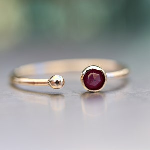 May include: A gold ring with a single red gemstone. The ring is open on one side and has a small ball on the other side.