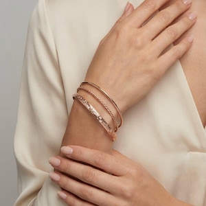 A person's arm wearing three rose gold bracelets: one engraved with a floral pattern, and two plain, thin bangles.