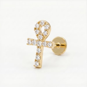 May include: A gold-colored ankh-shaped earring with clear, round gemstones. The earring has a post backing for pierced ears. The ankh symbol is a cross with a loop at the top, a symbol of life.