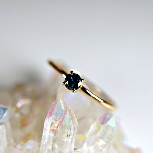 Puede incluir: Un anillo de oro con una sola piedra preciosa redonda de color azul oscuro engastada en un engaste de cuatro garras.