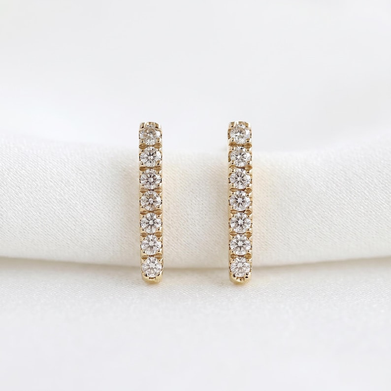 A pair of gold bar stud earrings embedded with small diamonds, standing upright on a soft white cloth.