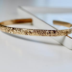 A single gold open cuff bracelet with a floral and swirl pattern engraved on the outside, resting on a white surface.