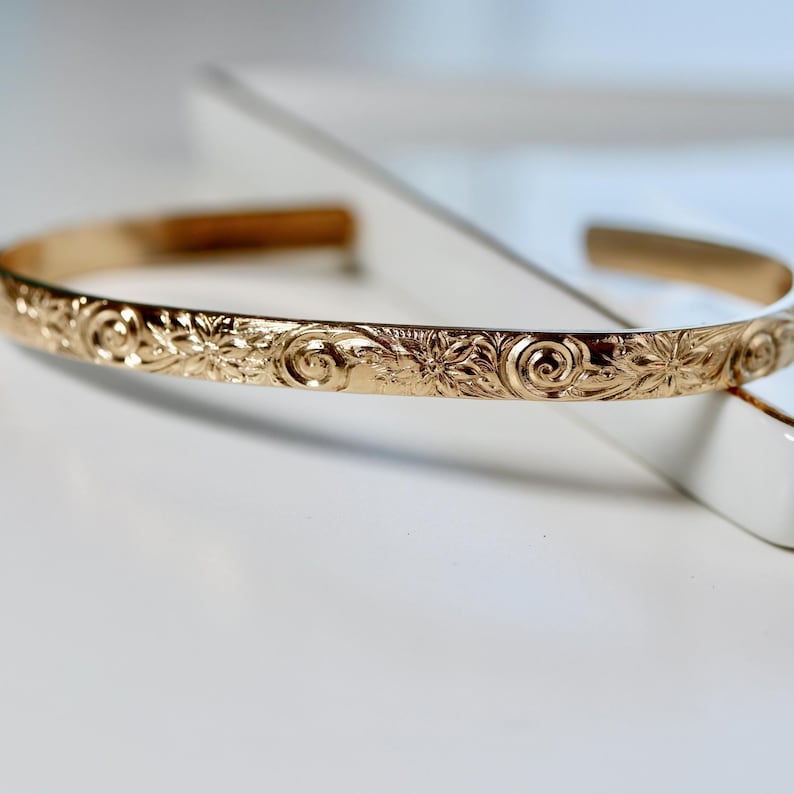 A single gold open cuff bracelet with a floral and swirl pattern engraved on the outside, resting on a white surface.