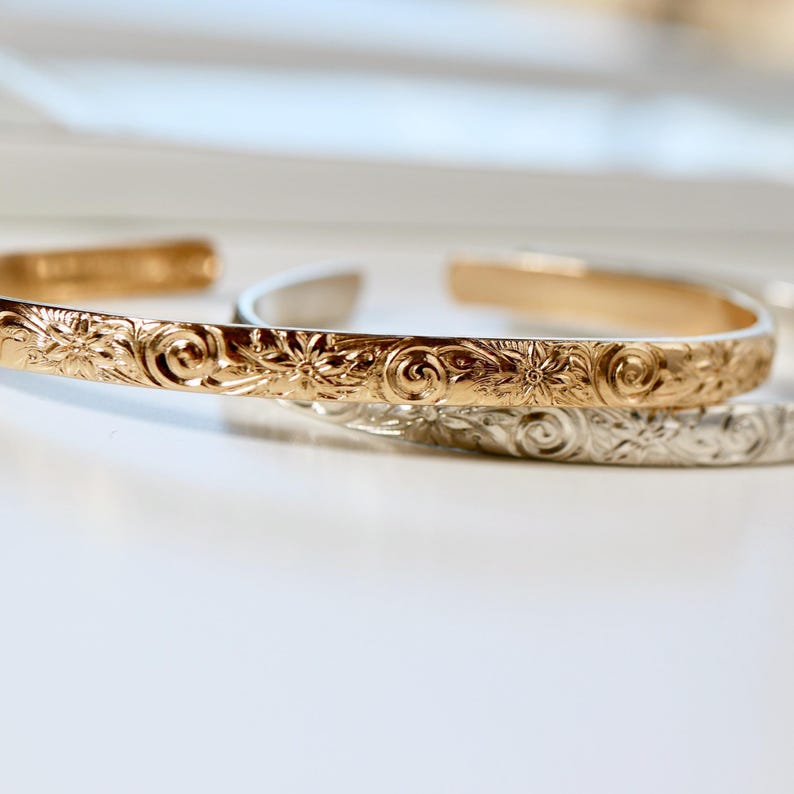 Three open cuff bracelets, two gold and one silver, with a floral and swirl pattern engraved on the outside.