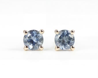 Puede incluir: Un par de pendientes de botón con piedras preciosas redondas de color azul claro engastadas en garras doradas. Los pendientes se muestran sobre un fondo blanco, resaltando el color y el diseño de la joyería.