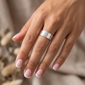 A wide, hammered silver band ring is worn on the ring finger of a person with manicured nails, against a blurred background of dried plants.