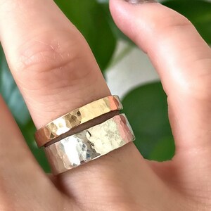 A wide, hammered silver band ring and a narrower, hammered gold band ring are stacked on a finger, with a green leaf in the blurred background.