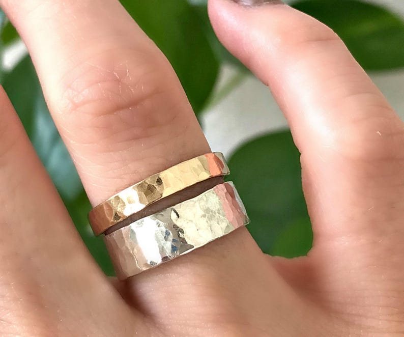 A wide, hammered silver band ring and a narrower, hammered gold band ring are stacked on a finger, with a green leaf in the blurred background.