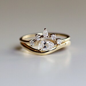 Pode incluir: Um anel de noivado de diamante em forma de p&ecirc;ra com uma banda delicada e uma coroa de diamantes menores. O anel &eacute; de ouro amarelo.