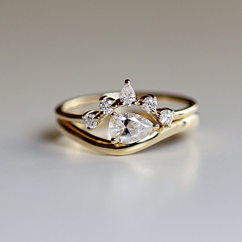 Pode incluir: Um anel de noivado de diamante em forma de p&ecirc;ra com uma banda delicada e uma coroa de diamantes menores. O anel &eacute; de ouro amarelo.