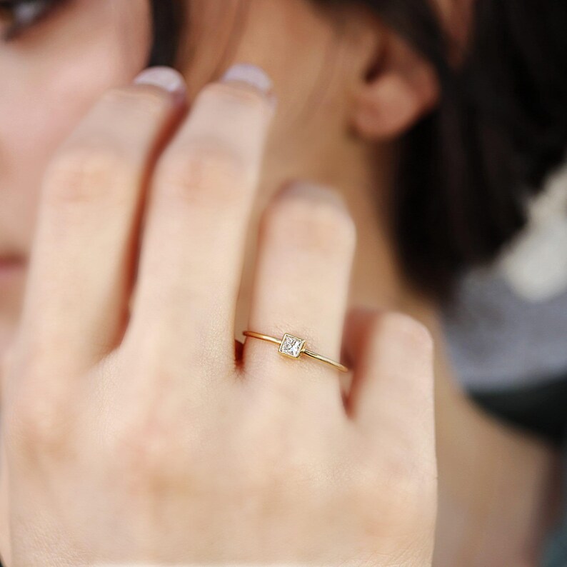 A close-up of a person wearing a slim gold ring with a small square diamond on their finger.