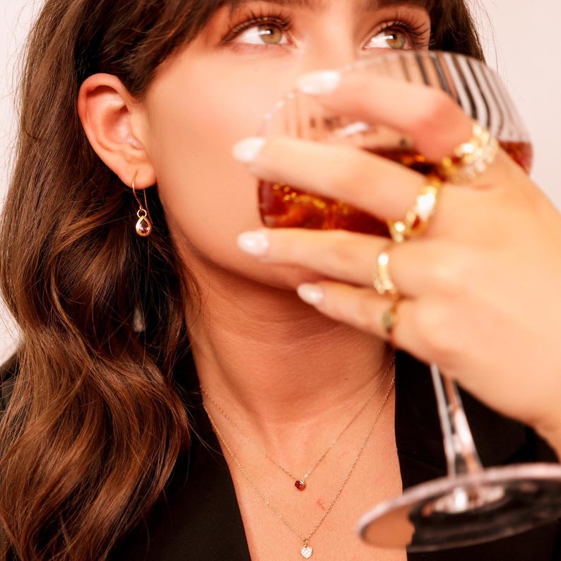 May include: A woman wearing a black blazer and gold jewelry, including a pair of teardrop earrings and two necklaces with a small heart and a small red stone pendant. She is holding a glass of amber liquid.