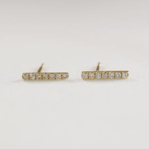 Two gold bar earrings featuring inlaid small diamonds, lying horizontally on a white textured background with posts visible.