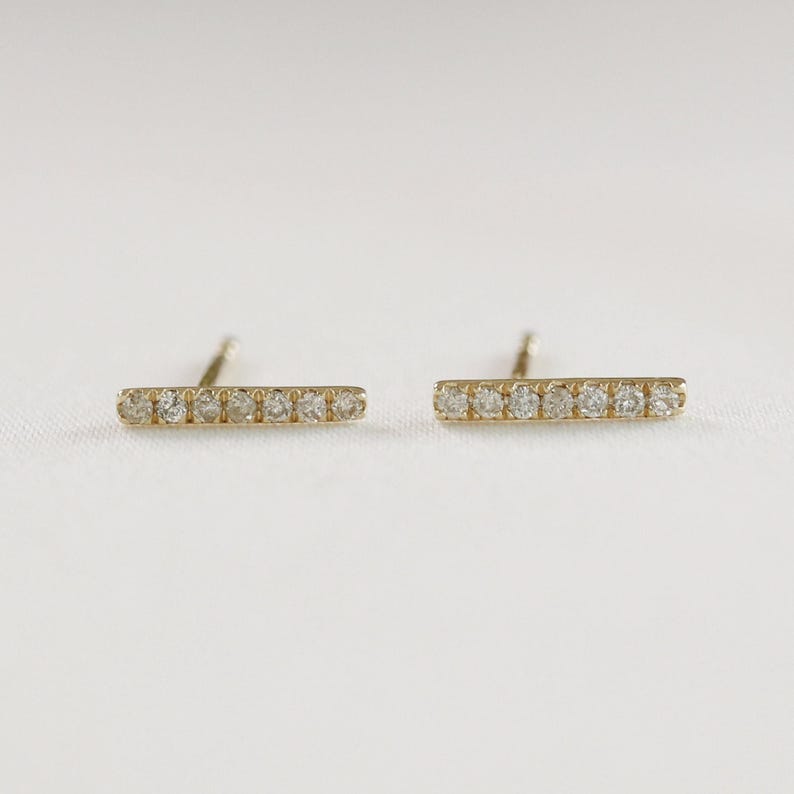 Two gold bar earrings featuring inlaid small diamonds, lying horizontally on a white textured background with posts visible.