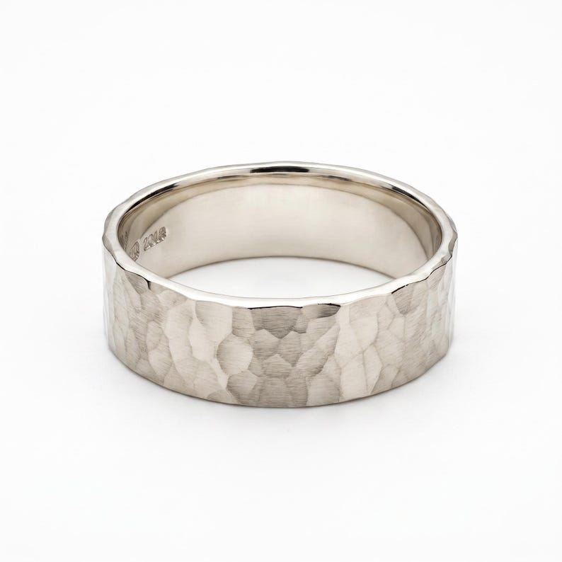 A single, wide, hammered silver band ring with a slightly irregular edge is shown on a white background.