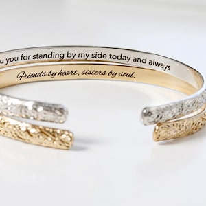 Two open cuff bracelets, one gold and one silver, with engraved text on the inside that reads: "Thank you for standing by my side today and always" and "Friends by heart, sisters by soul."